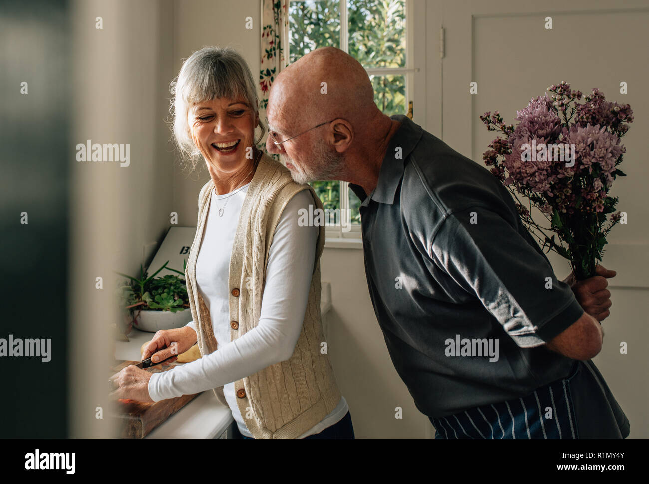 Senior uomo nascondere un mazzo di fiori dietro di lui a sorpresa la moglie. La donna come tagliare le verdure in cucina mentre il marito in piedi dietro di lei con un Foto Stock