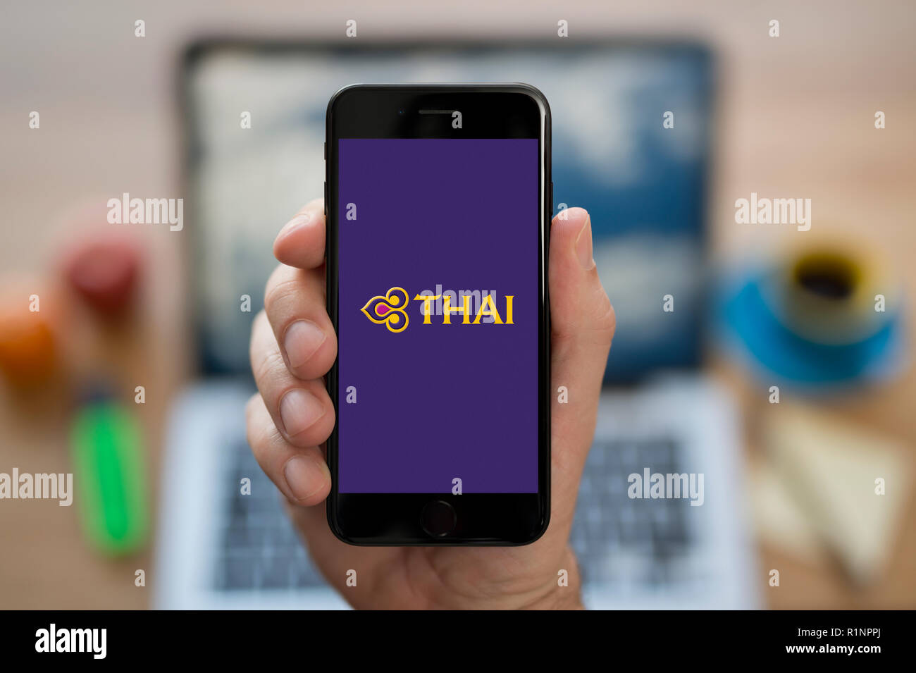 Un uomo guarda al suo iPhone che visualizza la Thai Airways logo, mentre se ne sta seduto alla sua scrivania per computer (solo uso editoriale). Foto Stock