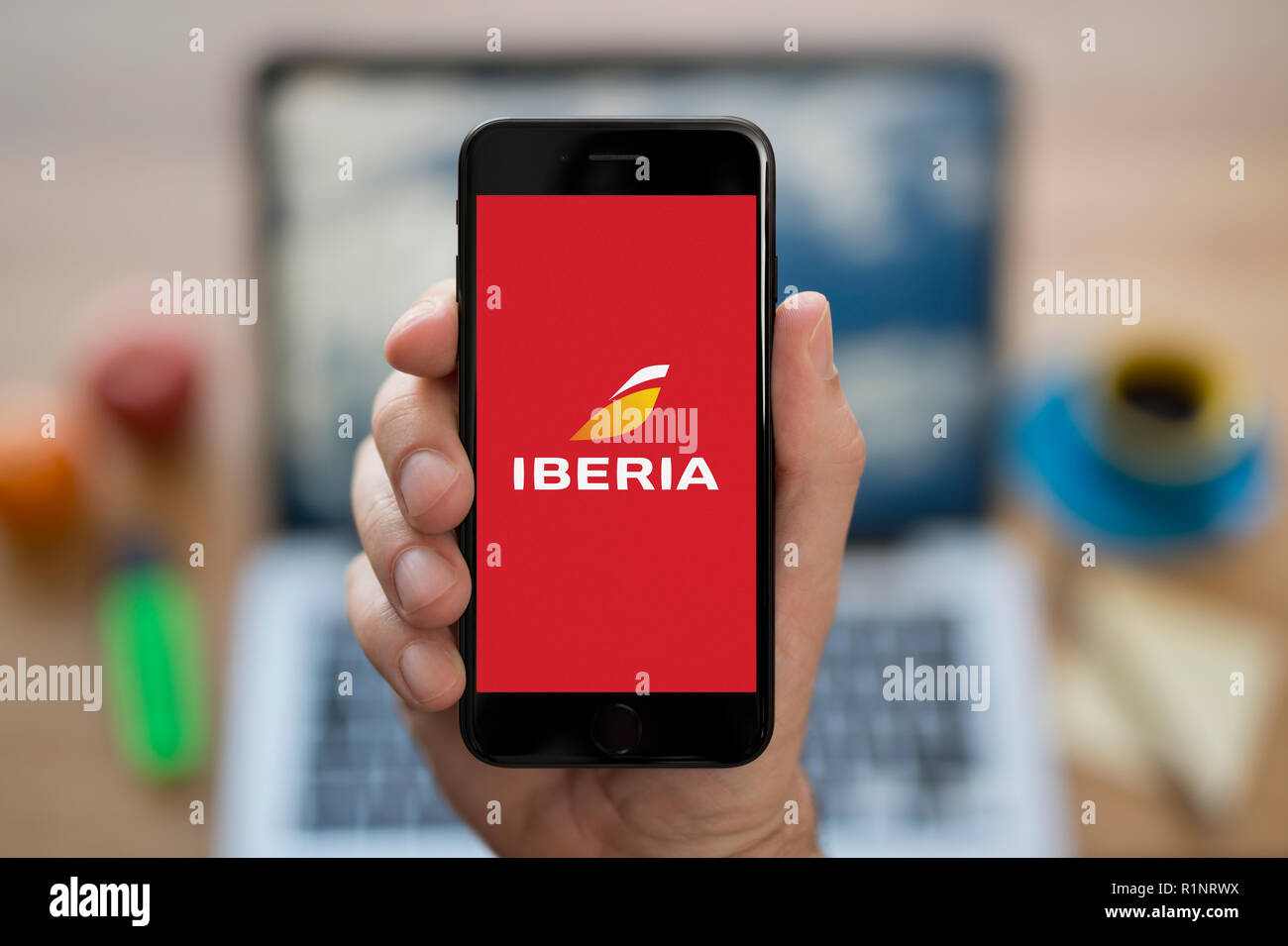 Un uomo guarda al suo iPhone che visualizza il logo di Iberia, mentre se ne sta seduto alla sua scrivania per computer (solo uso editoriale). Foto Stock