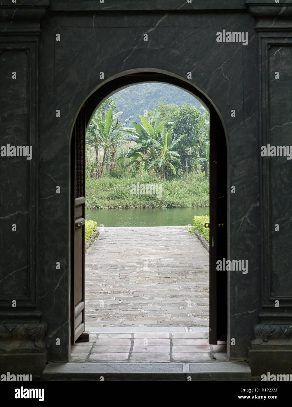 Ninh Binh, Vietnam. Foto di grigio di un portale in pietra che conduce a un giardino con acqua verde e alberi di banane. Foto Stock