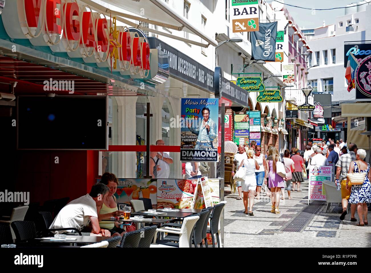I turisti in un momento di relax a un cafe' sul marciapiede nel centro storico della città con i suoi negozi e gli acquirenti al posteriore, Albufeira, Algarve, Portogallo, dell'Europa. Foto Stock