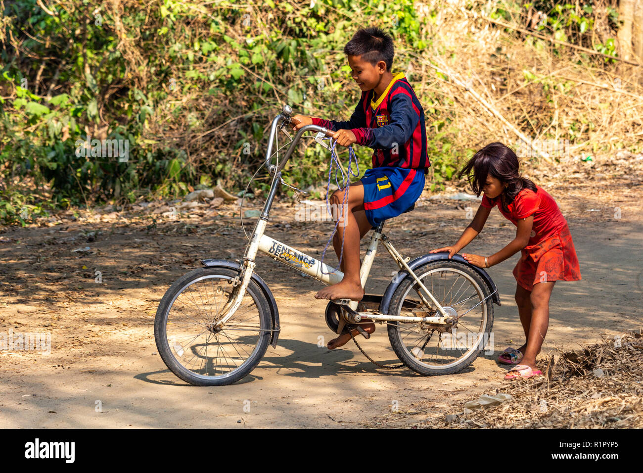Don Det, Laos - Aprile 22, 2018: Ragazza aiutando un ragazzo a guidare una bicicletta su un percorso in un remoto villaggio del sud Laos Foto Stock