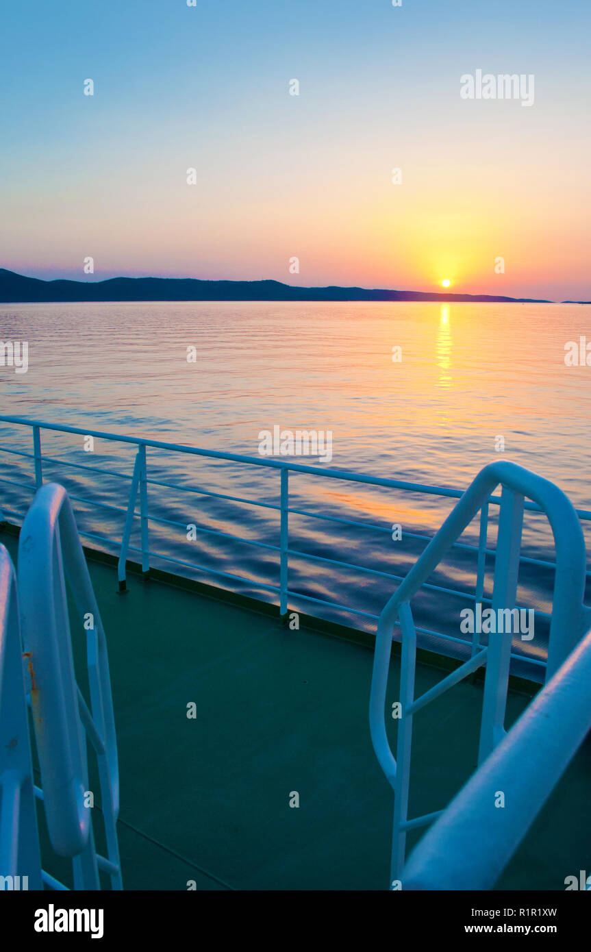 Impostazione del Sole sopra una collina gamma in mare in prossimità della costa della Croazia. Foto scattata dal ponte di una nave. ringhiere blu sulla parte anteriore Foto Stock
