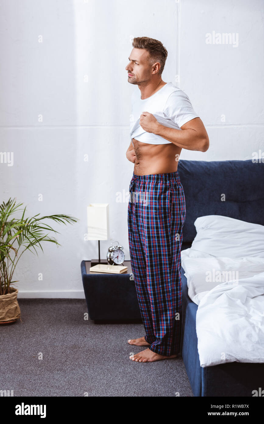 Uomo adulto mettendo su t-shirt bianco vicino al letto di casa Foto Stock