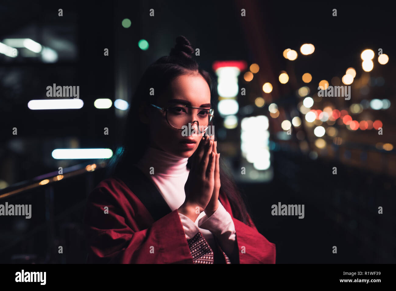 Bella ragazza asiatica in Borgogna kimono rendendo namaste gesto su strada con luci al neon in serata, città di future concept Foto Stock