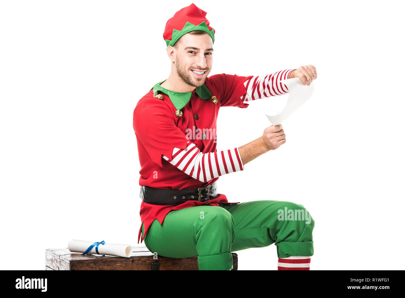 Uomo sorridente in elf natale costume seduto e azienda wishlist roll isolato su bianco Foto Stock