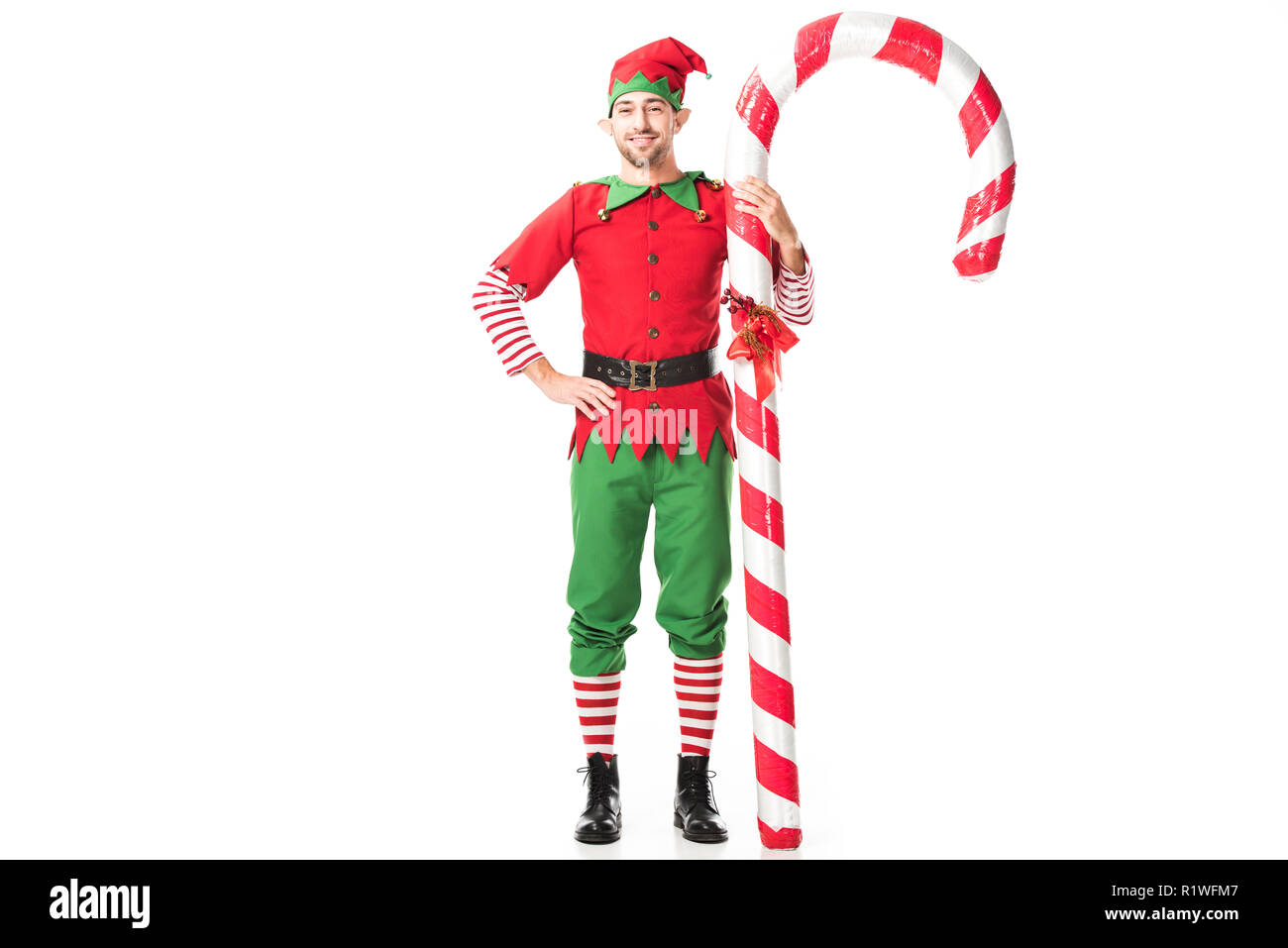Uomo sorridente in elf natale costume con mani sui fianchi in piedi vicino al grande candy cane isolato su bianco Foto Stock