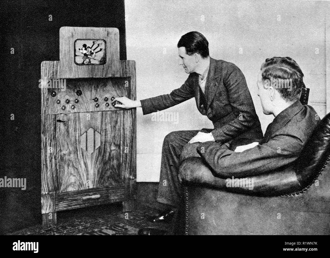 Un 1935 fotografia che mostra i primi giorni della televisione in Gran Bretagna - due uomini in abiti di sedersi a guardare Mickey Mouse riproduzione di cornamuse sul loro piccolo televisore alloggiato all' interno di un grande armadio in legno Foto Stock