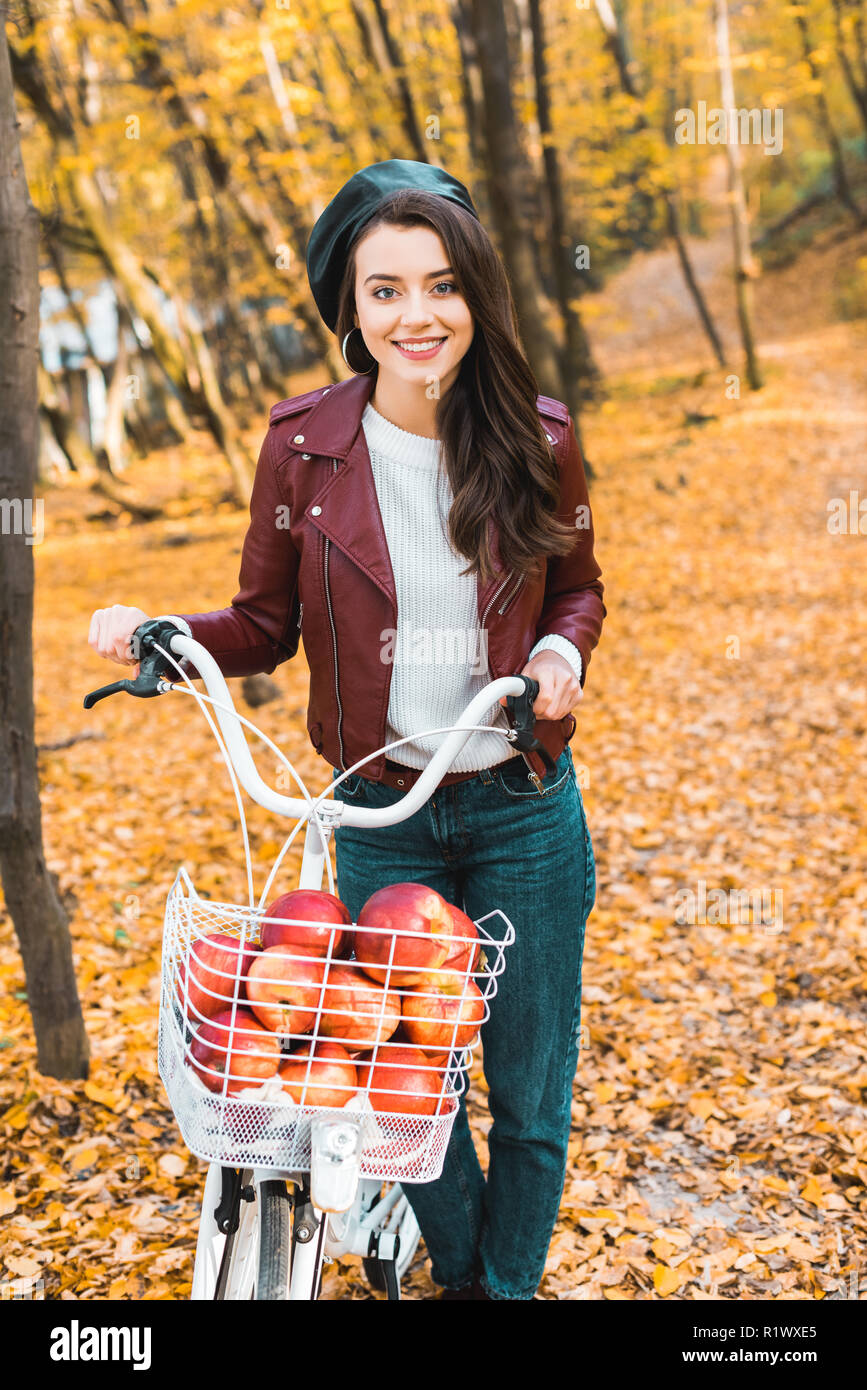 Sorridente ragazza elegante in giacca di pelle che porta bicicletta con cesto pieno di mele rosse nel parco autunnali Foto Stock