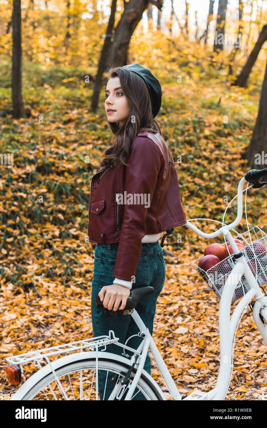 Attraente ragazza elegante in berretto e giacca di pelle in posa vicino a bicicletta in foresta autunnale Foto Stock