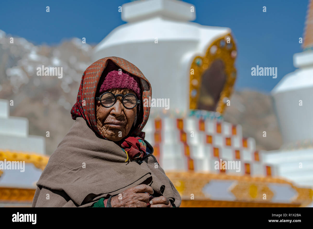 Un ritratto di una vecchia donna locale, indossando occhiali, stupa buddisti e montagne coperte di neve in background Foto Stock