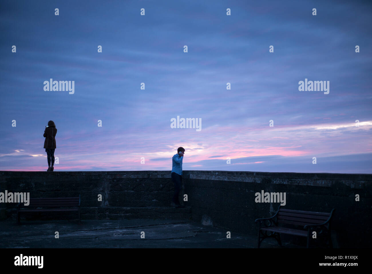 Giovane donna prende uno smartphone fotografia di colori pastello del tramonto mentre il suo partner prende una chiamata sul suo smartphone, Galles Foto Stock