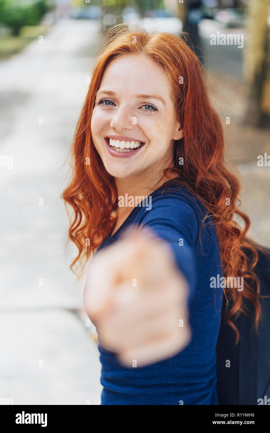 Attraente redhead donna puntando verso la telecamera con un sorriso vivace in una strada urbana con particolare attenzione per il suo volto Foto Stock