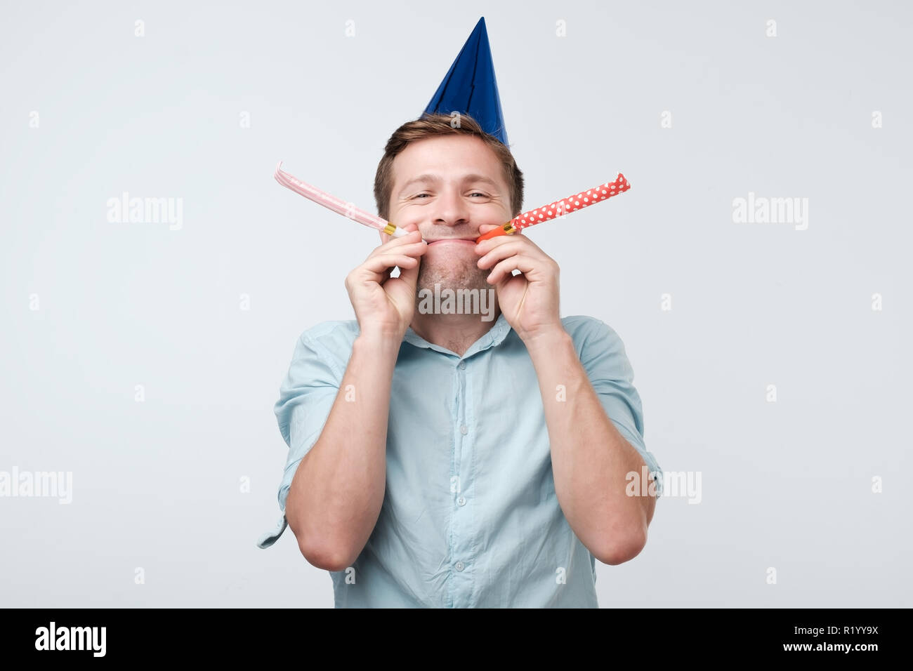 Bello allegro europeo del giovane uomo divertirsi su un partito che indossa blu denim shirt e vacanze hat, partito di soffiaggio avvisatore acustico. Foto Stock