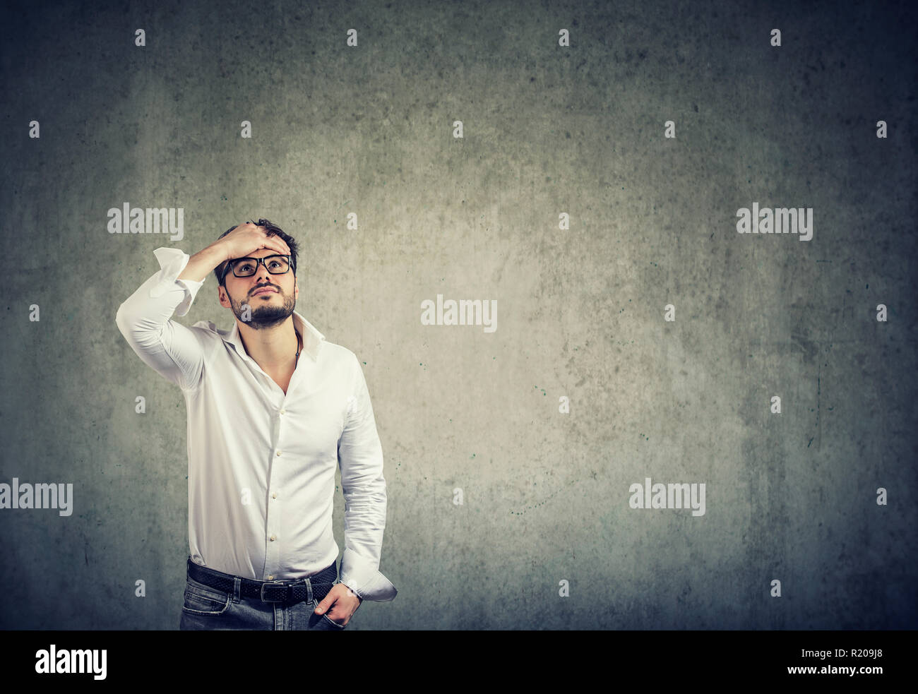 Giovane uomo informale in una camicia bianca e occhiali aventi problemi e cercare nella sfida sul muro grigio sfondo Foto Stock