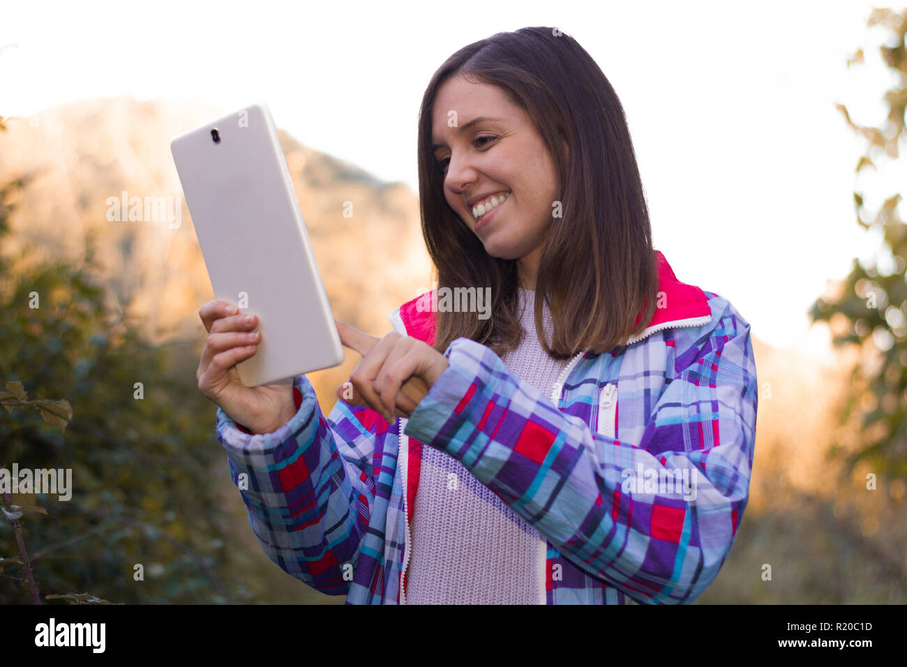 Divertente e sorridente giovane donna che interagisce con il suo tablet in natura e montagna cercando la migliore offerta Foto Stock