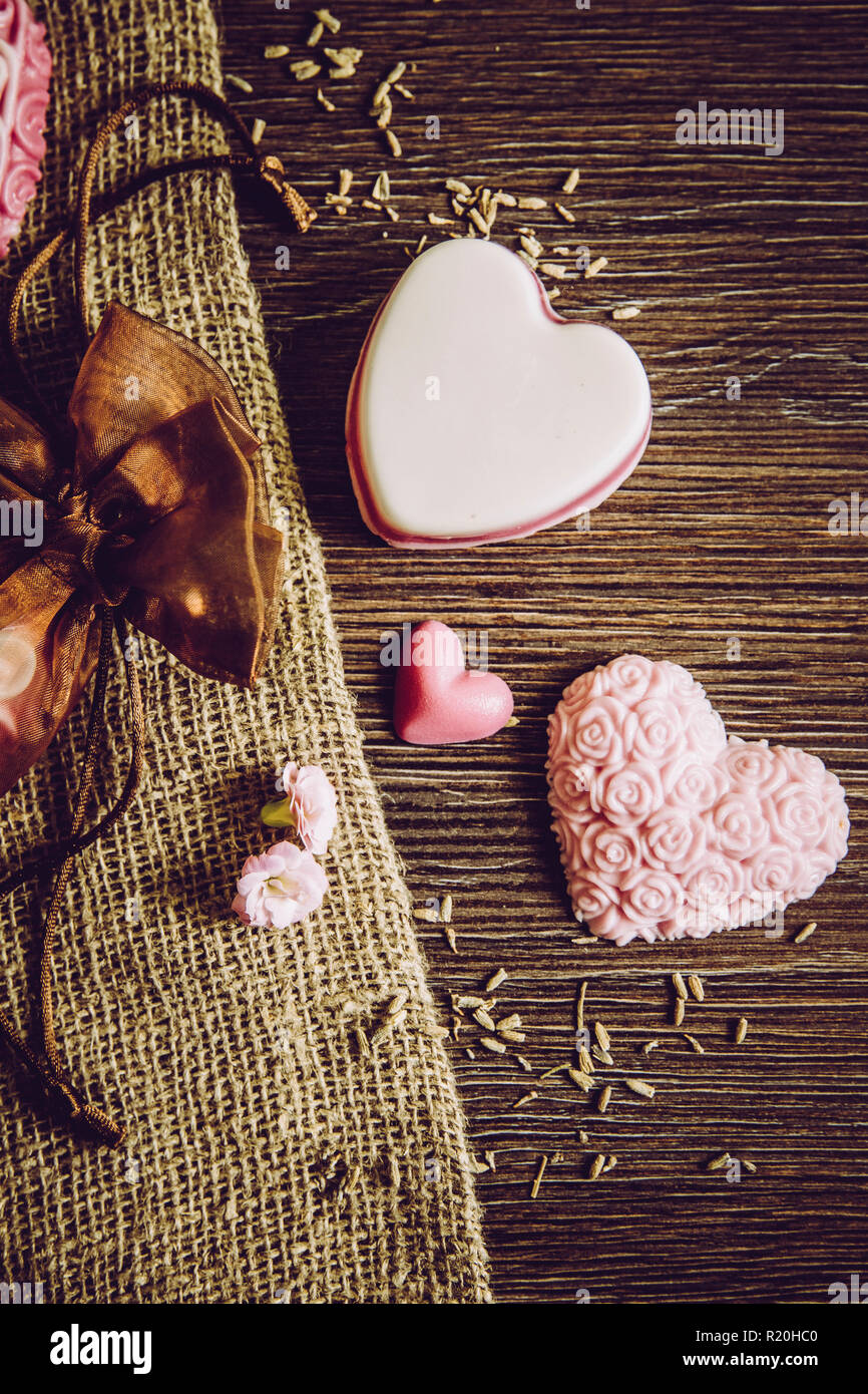 I diversi colori fatto a mano forma di cuore saponi sulla tavola di legno sfondo, tela ruvida stoffa e in tulle marrone borsa regalo. Fatto a mano idea regalo concetto. Sacco di co Foto Stock
