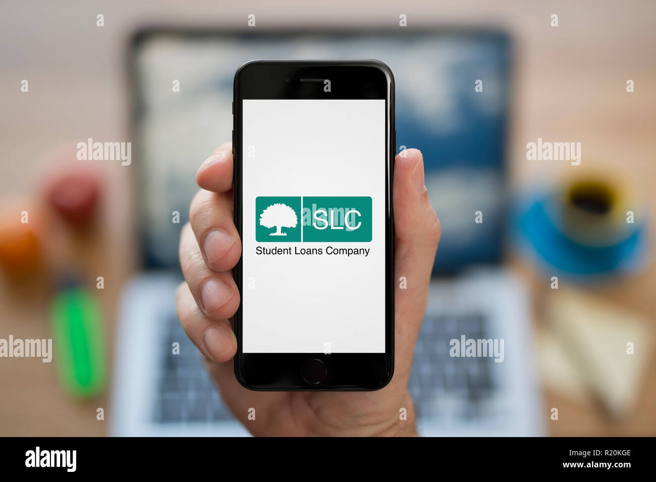 Un uomo guarda al suo iPhone che visualizza la Student Loans Company (SLC) il logo, mentre se ne sta seduto alla sua scrivania per computer (solo uso editoriale). Foto Stock