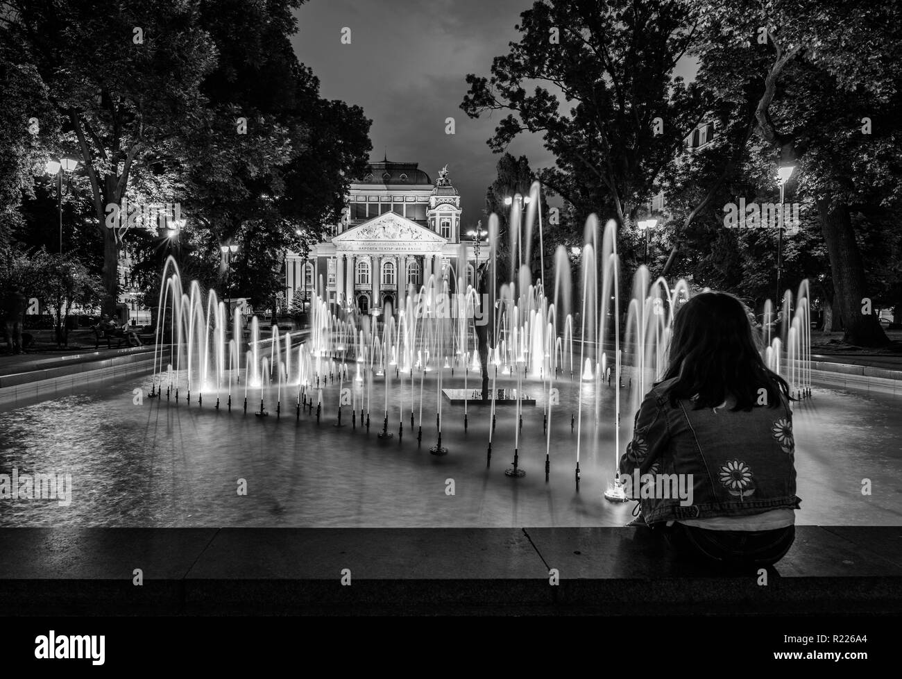 Giovane ragazza seduta da una fontana di notte, guardando il palazzo del Teatro Nazionale - bella tenebrologo, luci della città Foto Stock