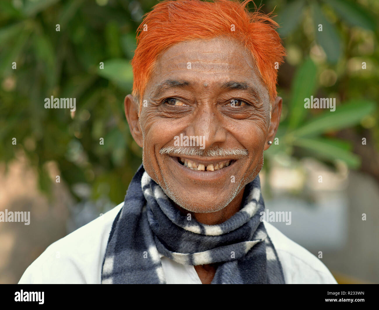 Strizzare gli occhi, di mezza età uomo indiano (occhio destro deviato verso l'esterno) con una matita di baffi e henné-tinto arancio sorrisi dei capelli per la fotocamera. Foto Stock