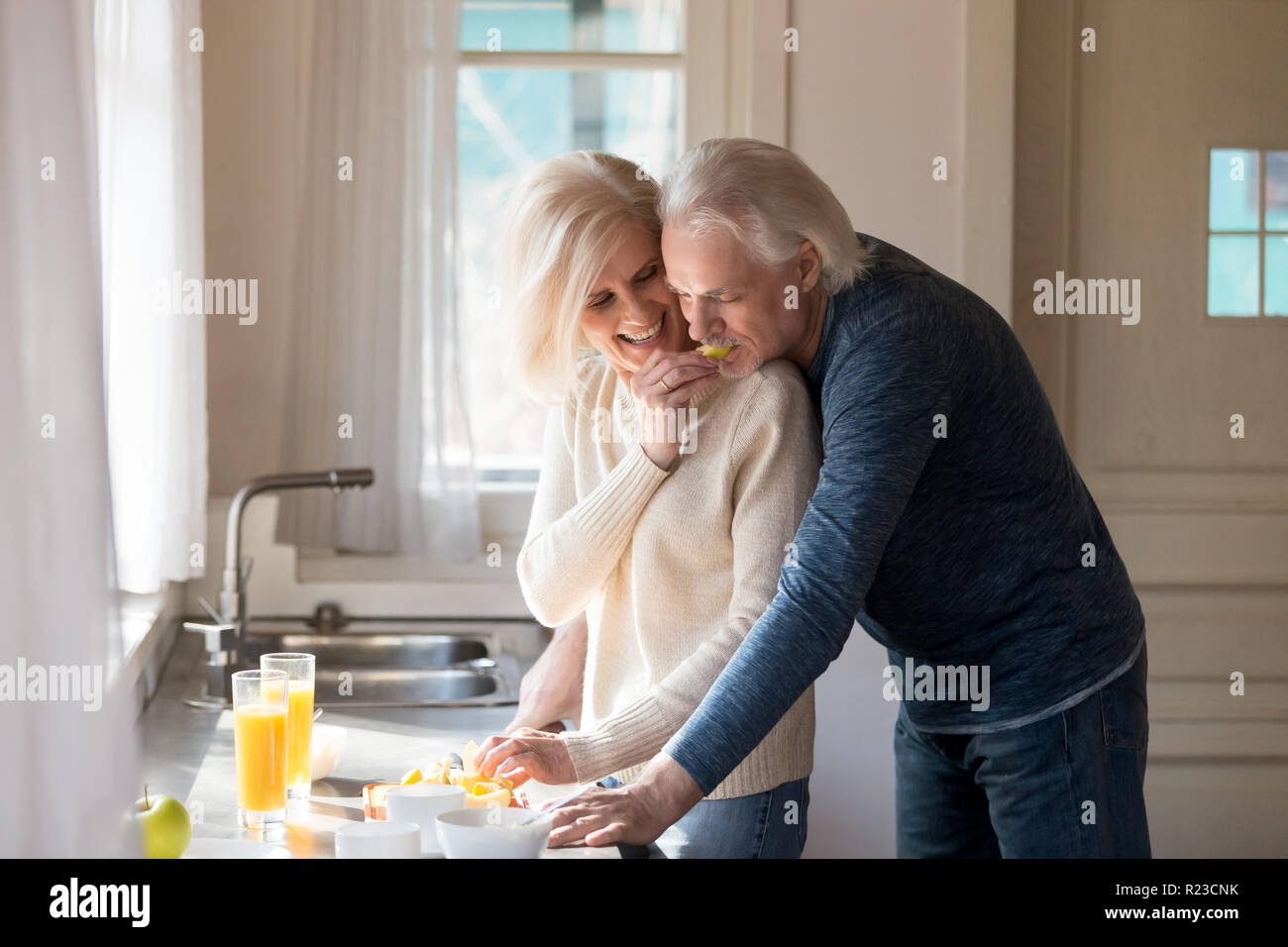 Senior marito amorevole abbraccio moglie da dietro la preparazione di cibo in cucina, caring sorridente di età della donna amata alimentazione uomo mentre la cottura a casa, coppia di anziani Foto Stock