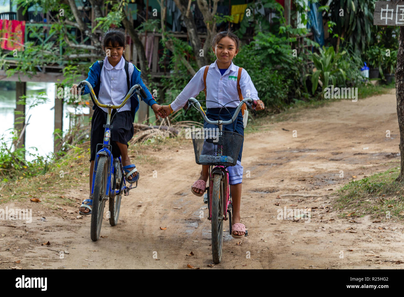 Don Det, Laos - Aprile 24, 2018: scuola locale ragazze tenendo le mani mentre si guida biciclette su una strada fangosa Foto Stock