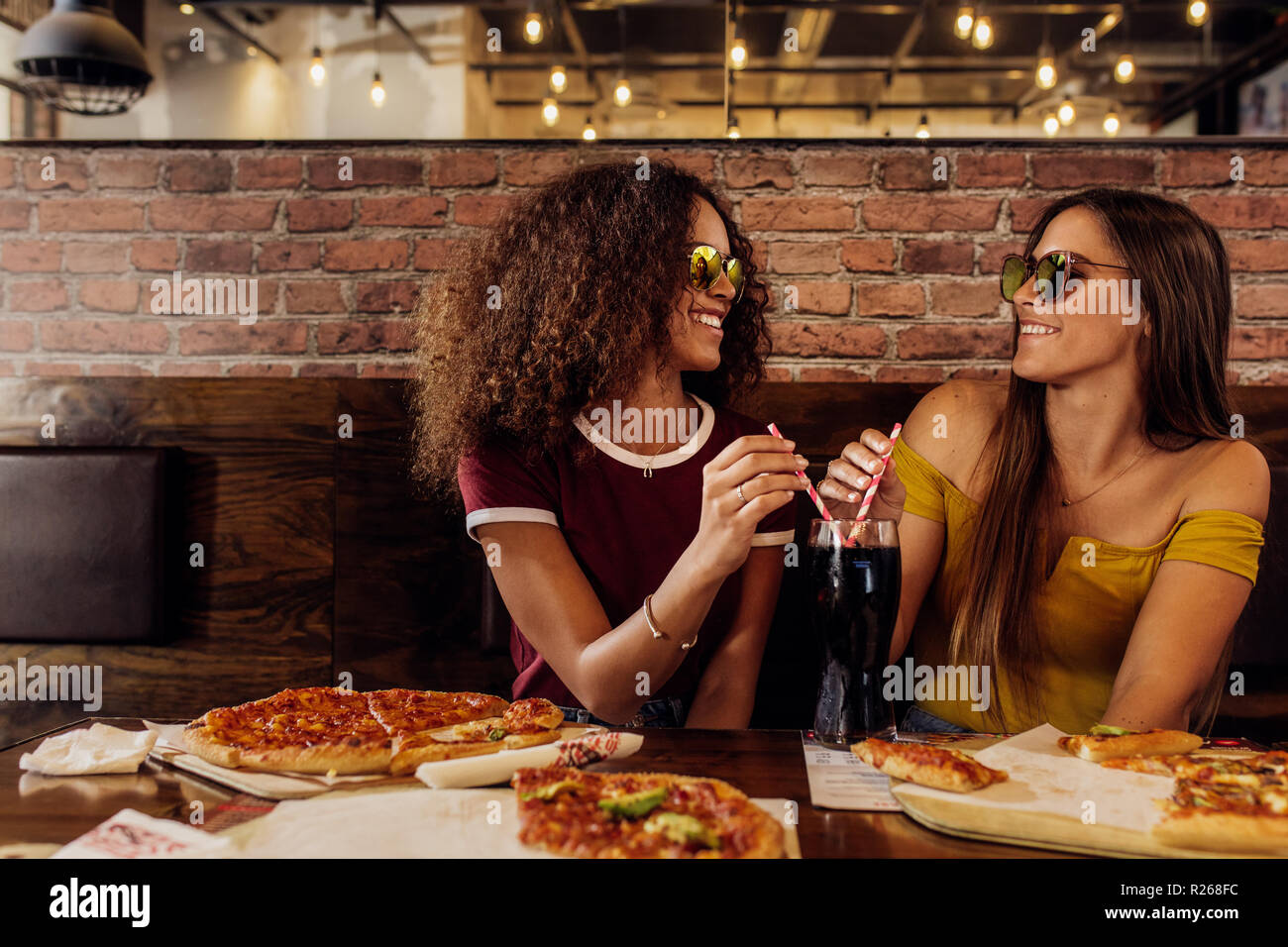 Elegante donne amici indossando occhiali da sole seduti ad un ristorante fast food. Due femmine di bere drink freddo presso il cafe con pizza sul tavolo. Foto Stock