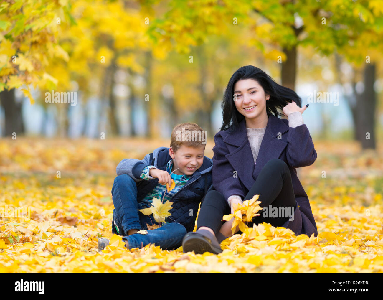 Un felice, contenuto madre e figlio seduti e giocando in un parco della città tra giallo Foglie di autunno. Foto Stock