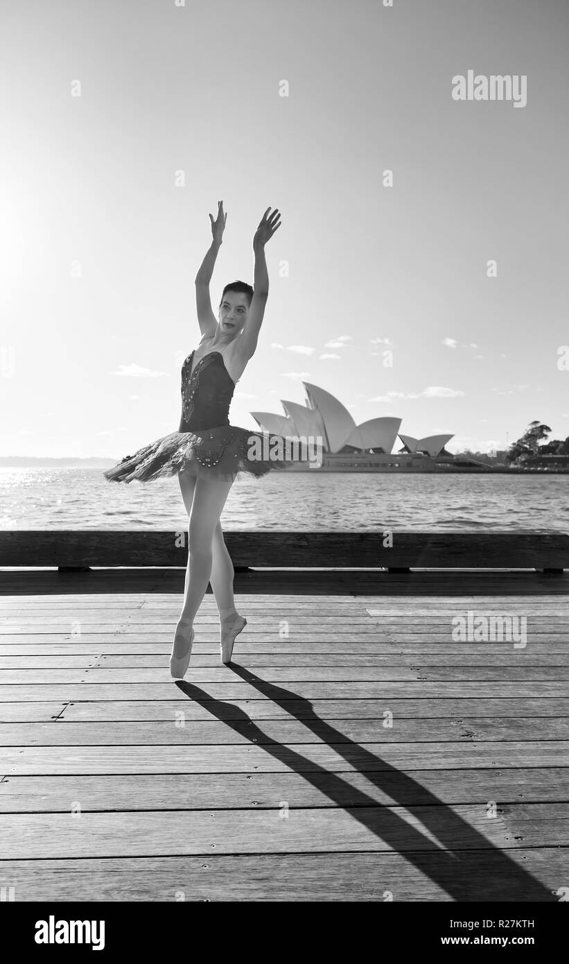 Montare giovane ballerina in rosso tutu ballando sul ponte di legno sulle rive del porto di Sydney in morbida luce mattutina in bianco e nero ad alto contrasto dell'umore. Foto Stock