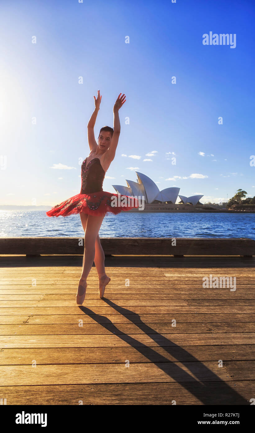 Montare giovane ballerina in rosso tutu ballando sul ponte di legno sulle rive del porto di Sydney in morbida luce mattutina sotto il cielo blu. Foto Stock