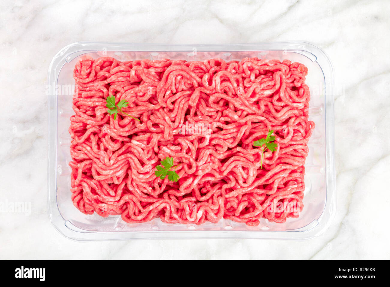 Una foto di carni macinate in un vassoio di plastica, ripresa dall'alto su un marmo bianco sfondo con foglie di prezzemolo Foto Stock