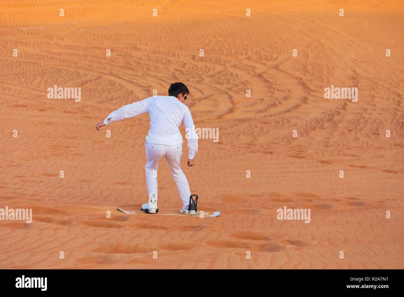 Giovane turista uomo vestito di bianco vestiti sand boarding giù dune di sabbia in un deserto vicino a Dubai, Emirati arabi uniti Foto Stock