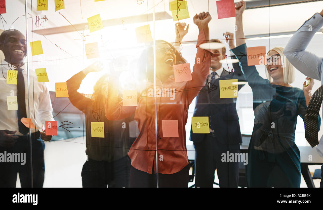 Gruppo estatica di diversi imprenditori urlando e saltando su e giù mentre il brainstorming con foglietti adesivi su una parete di vetro in un ufficio moderno Foto Stock