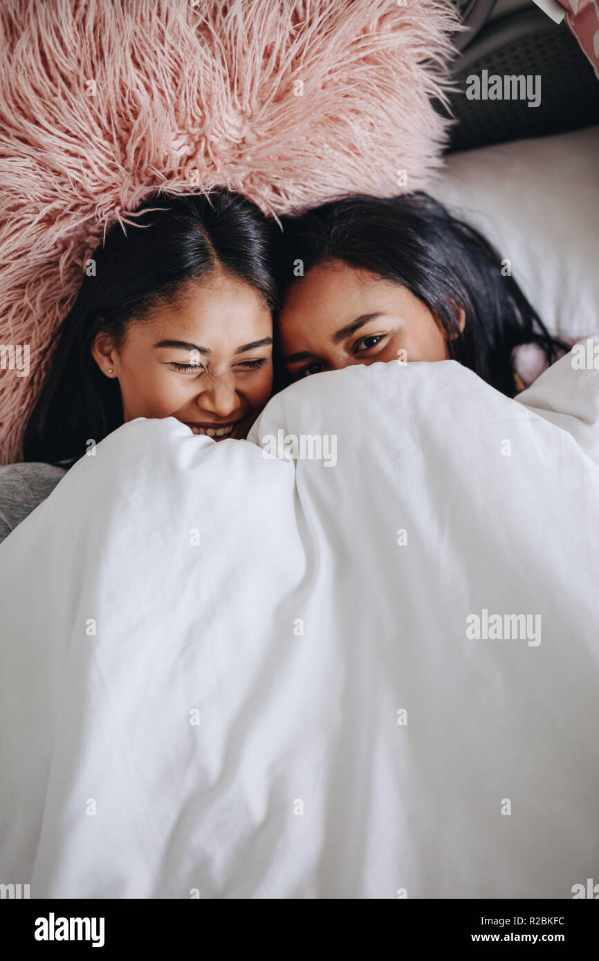 Vista dall'alto di ragazze giacente sul letto a copertura di una coperta e ridere. Due amiche ridere giacente sul letto durante un sleepover. Foto Stock