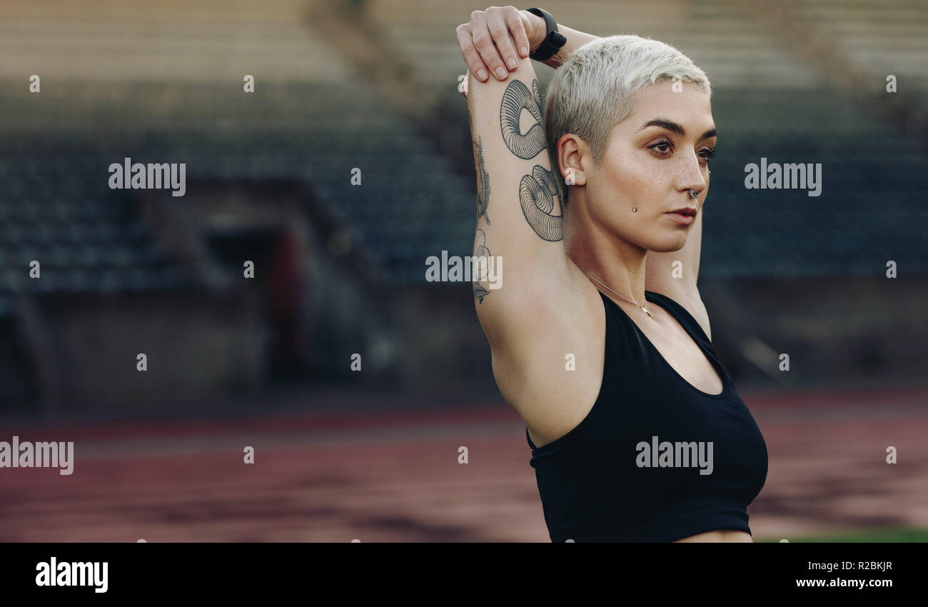 Vista laterale verticale di una femmina di runner facendo esercizi di stretching in piedi in una traccia e field stadium. Donna in abiti fitness stretching le sue braccia s Foto Stock