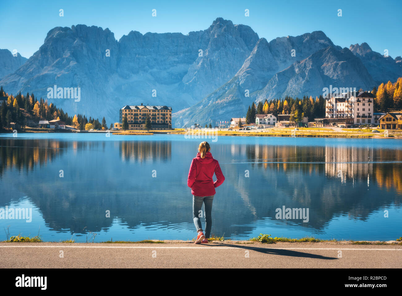 Giovane donna in piedi sulla costa del lago di Misurina a sunrise in autunno. Dolomiti, Italia. Paesaggio con ragazza in giacca rossa e la riflessione in acqua, bui Foto Stock