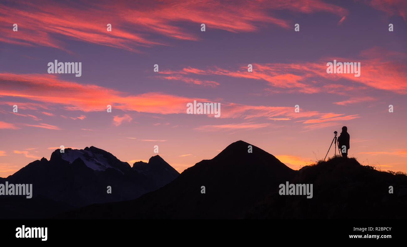 Silhouette di un fotografo sul picco di montagna contro la roccia e cielo viola con le Nuvole rosa al tramonto. Paesaggio con uomo, montagna cresta e colo Foto Stock