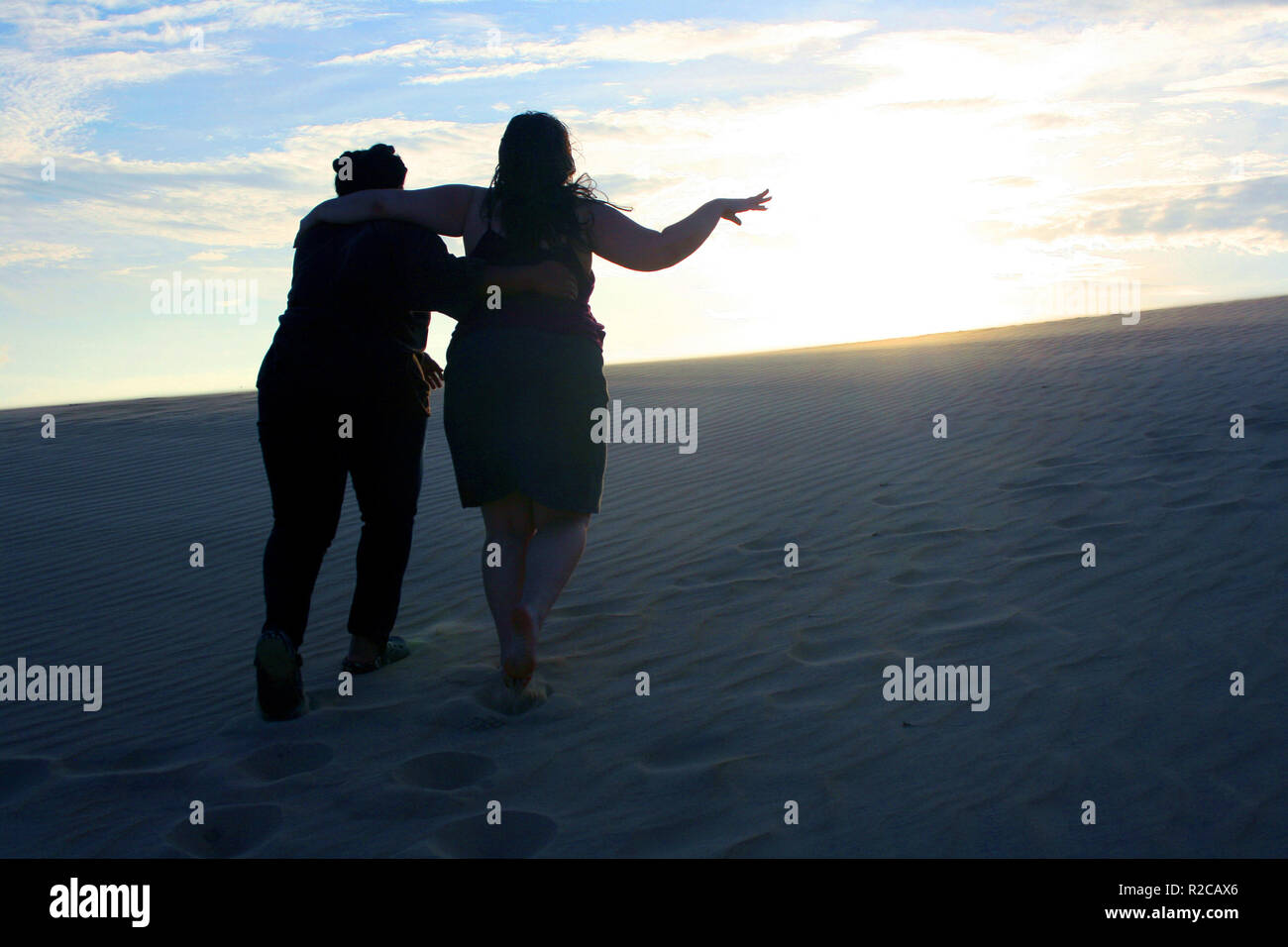 Due donne con le braccia intorno a ogni altro di cintura di salire un gigante di dune di sabbia verso il tramonto a Jockey Crinale del Parco statale in Kill Devil Hills, NC, Stati Uniti d'America Foto Stock