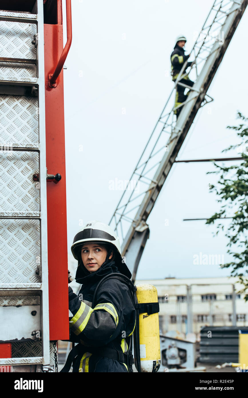 Firefighter femmina in uniforme e casco che guarda lontano mentre si collega in piedi sulla scala sulla strada Foto Stock