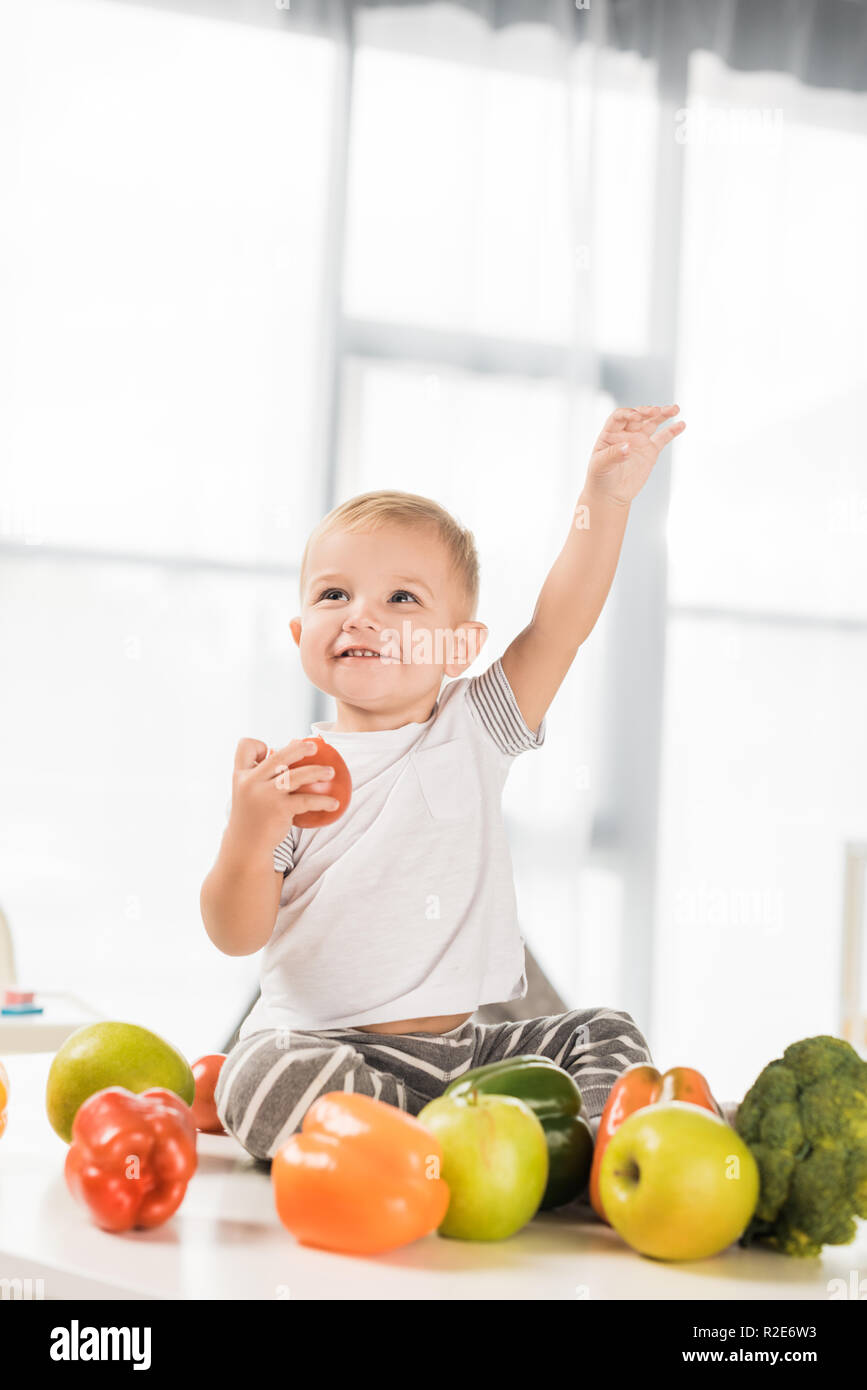Carino il toddler alzando la mano e seduta sul tavolo circondato da frutta e verdura Foto Stock