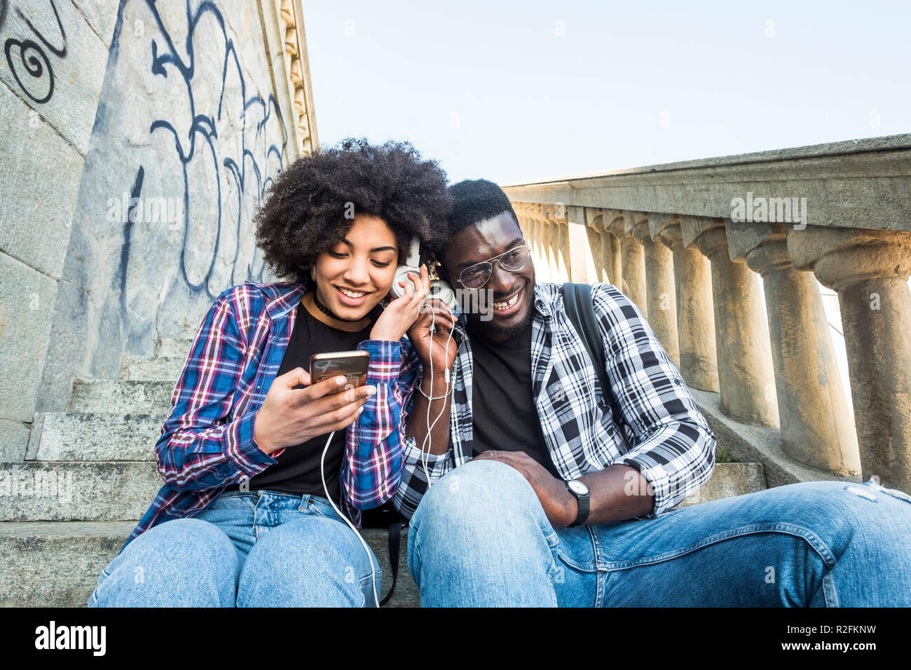 Allegro felice africann nero giovane seduto togethe in amicizia ascoltando la musica da un telefono moderno e condividendo le cuffie come partner. sorridente e divertirsi Foto Stock