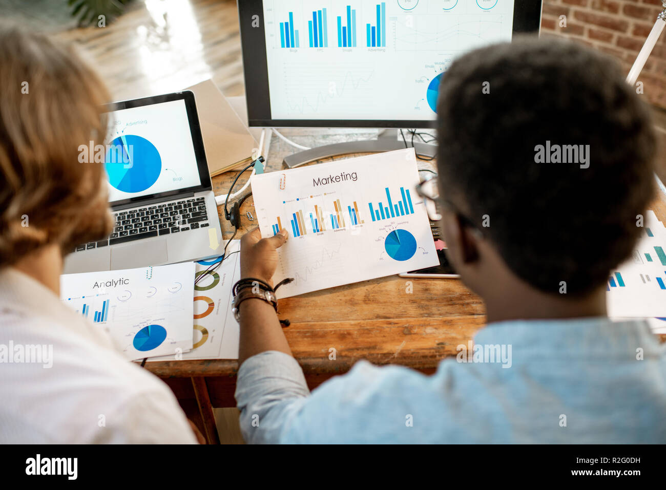 Due maschi collaboratori lavora con analitycs seduti insieme al posto di lavoro con computer, laptop e documenti. Vista da dietro incentrate sui documenti Foto Stock
