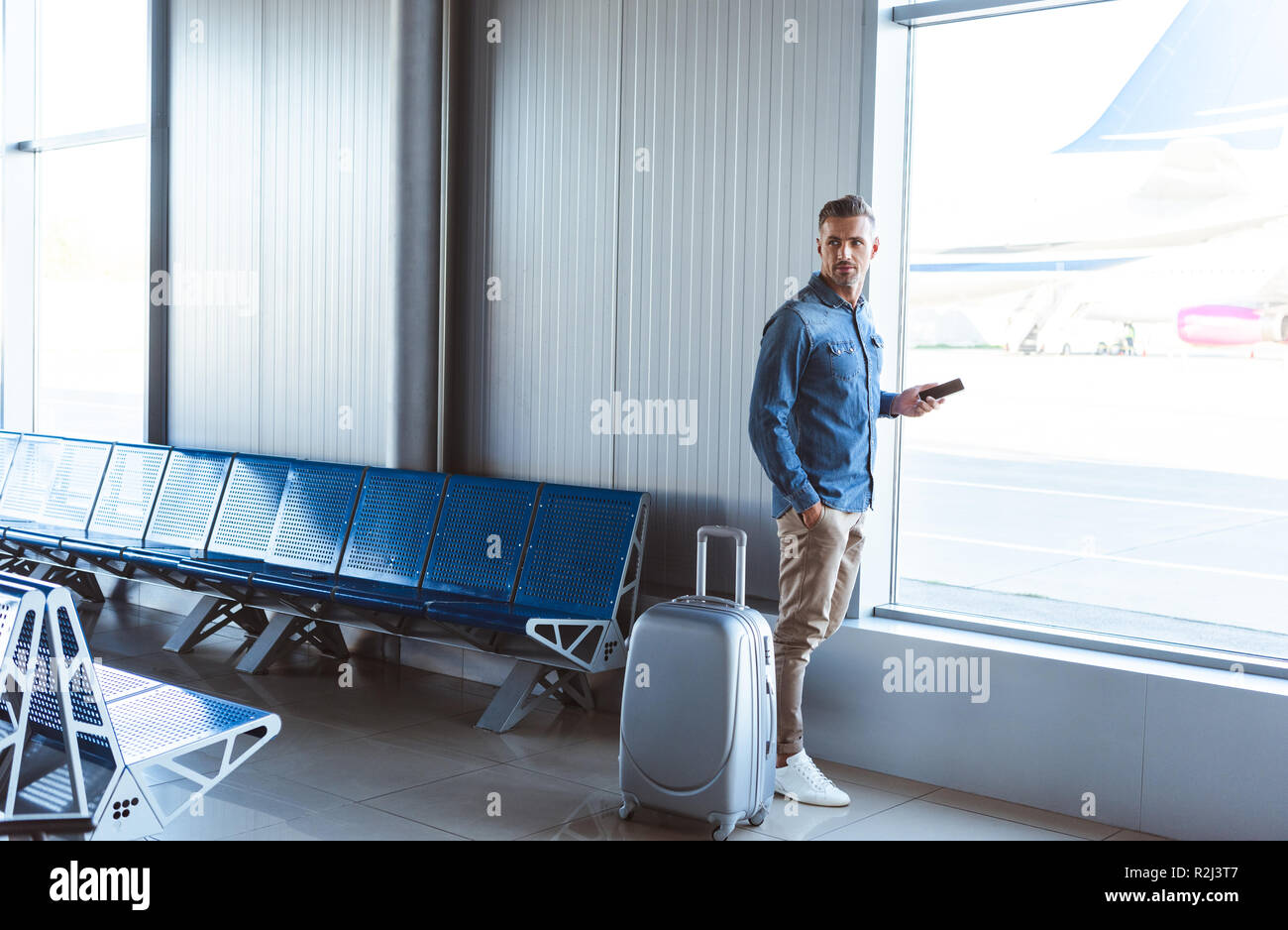 Uomo con argento Valigia controllo telefono e in attesa vicino alla finestra in aeroporto Foto Stock