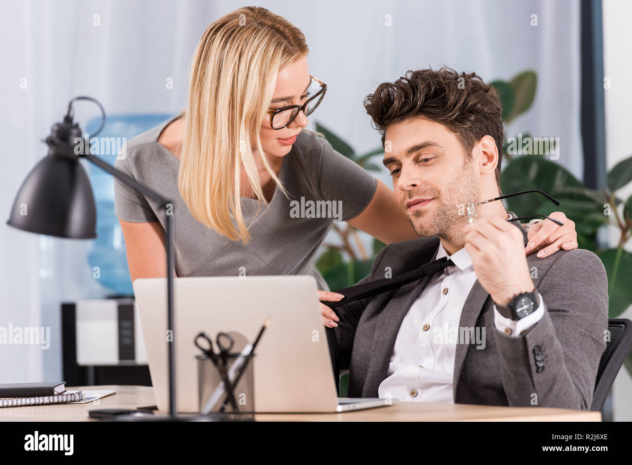 Ritratto di imprenditrice tirando i colleghi cravatta al lavoro con il computer portatile in ufficio, flirtare e ufficio concetto romantico Foto Stock