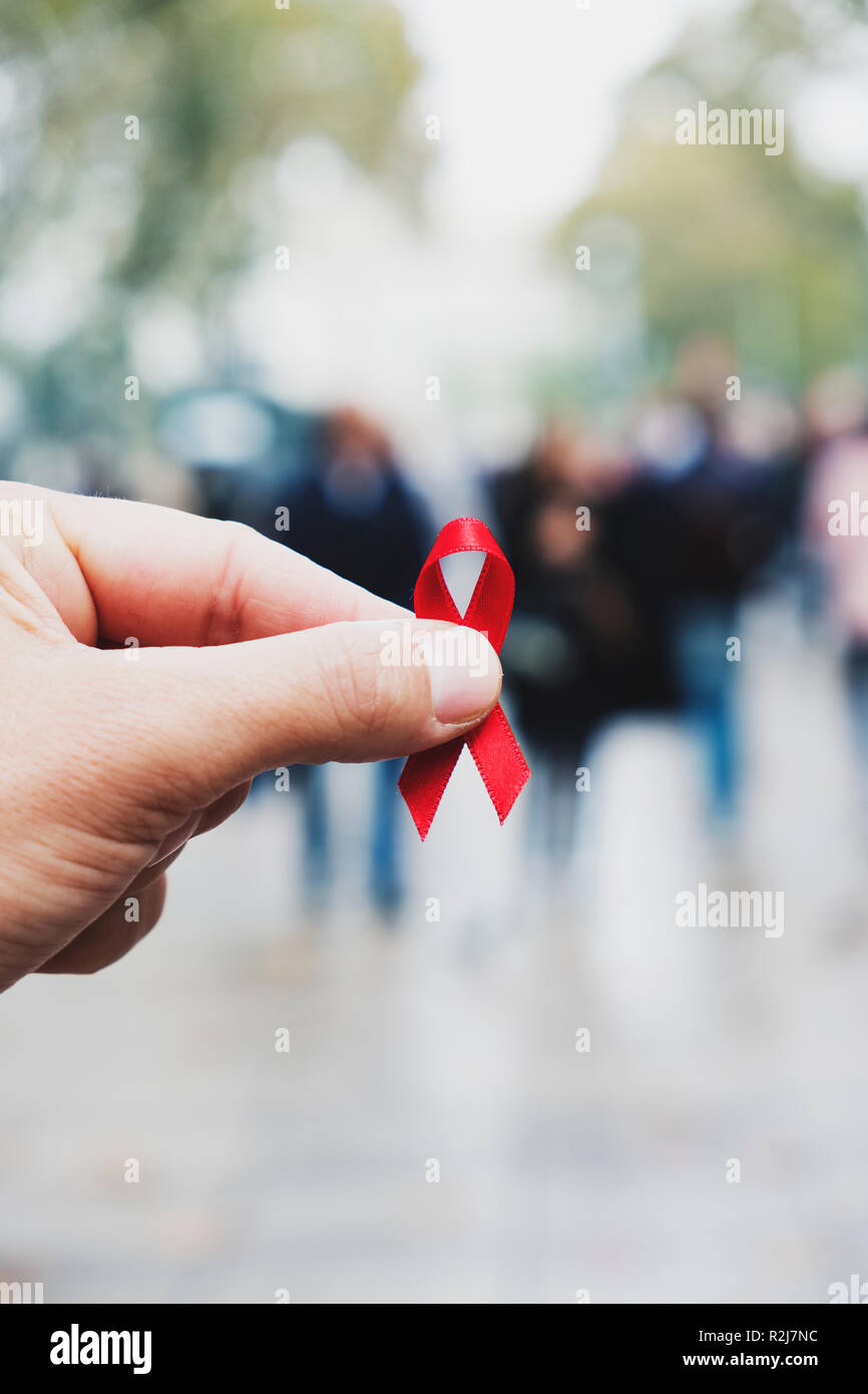 Primo piano di un red ribbon di sensibilizzazione per la lotta contro l AIDS in mano di un giovane uomo caucasico in una trafficata strada pedonale di una città Foto Stock