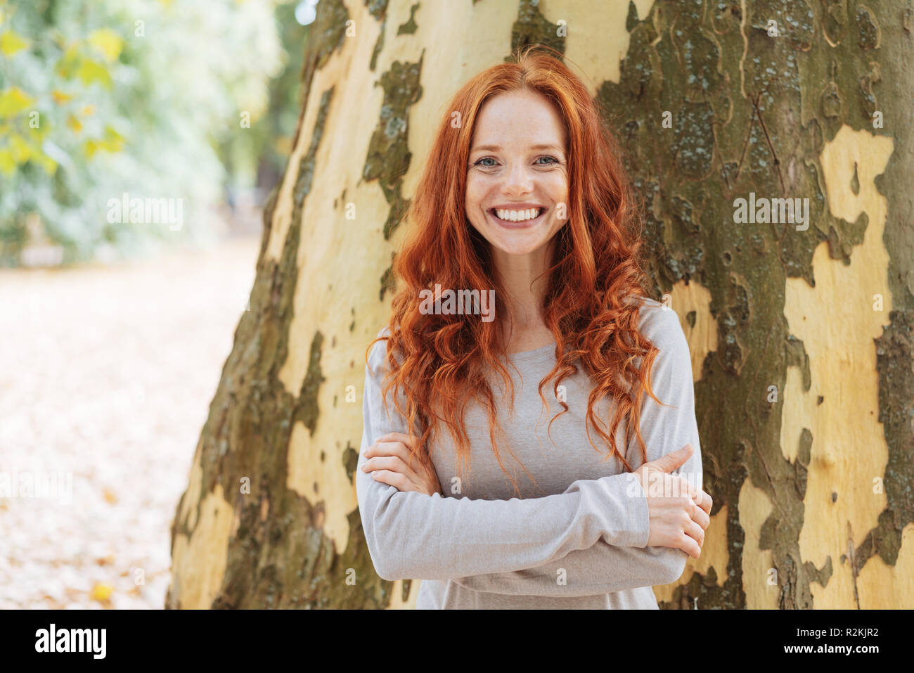 Sorridente fiducioso giovane donna con bella lunga parentesi capelli rossi in piedi con le braccia incrociate nella parte anteriore del tronco di un albero in un parco Foto Stock