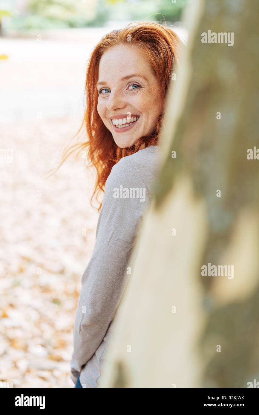 Carino carismatico redhead giovane donna appoggiata contro un albero girando a guardare indietro oltre il tronco alla fotocamera con un felice sorriso amichevole Foto Stock