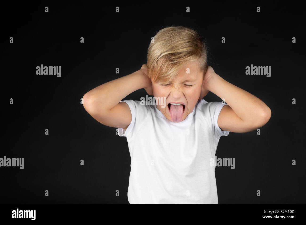 Piccolo ragazzo biondo copre le sue orecchie e bastoni fuori la sua lingua Foto Stock