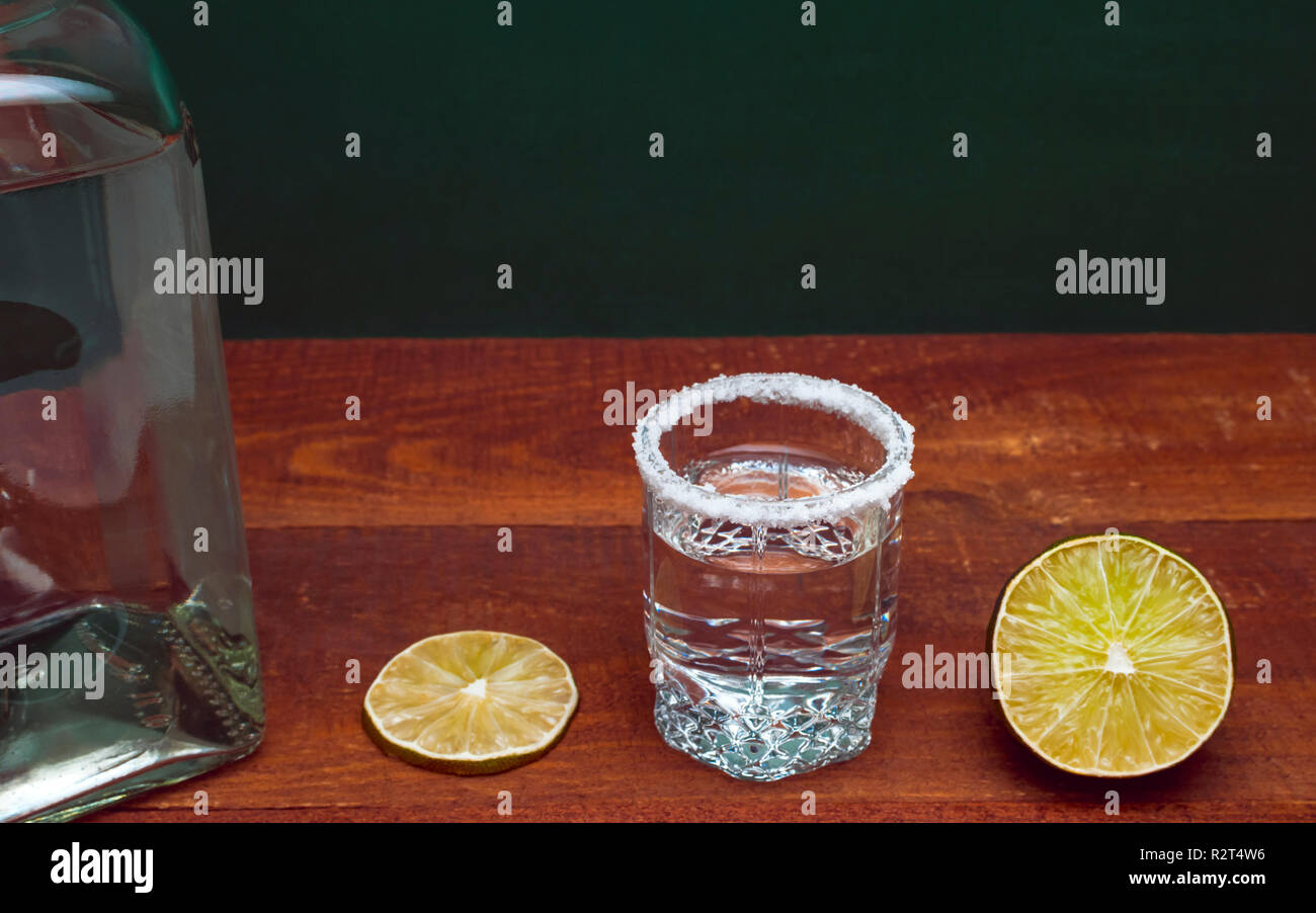 Argento alcool Tequila Shot bordata con sale su una tavola di legno tavolo rustico. Sezione trasversale di un lime, una fetta di lime e una bottiglia. Sfondo verde. Foto Stock