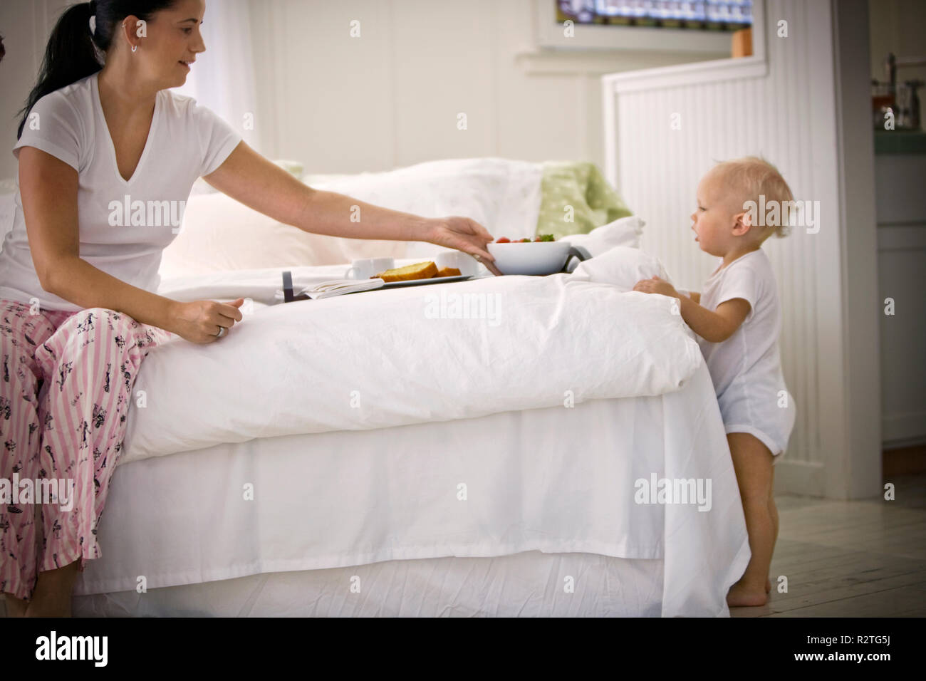 Mid-adulto donna seduta su di un letto con colazione mentre suo figlio toddler guarda a. Foto Stock